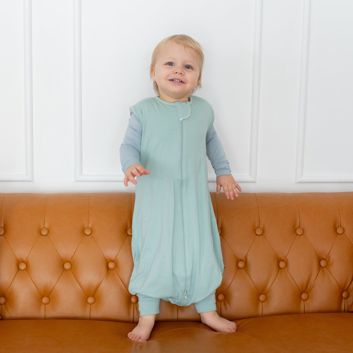 Toddler wearing Kyte Baby Sleep Bag Walker in Sage 0.5 while standing on a couch