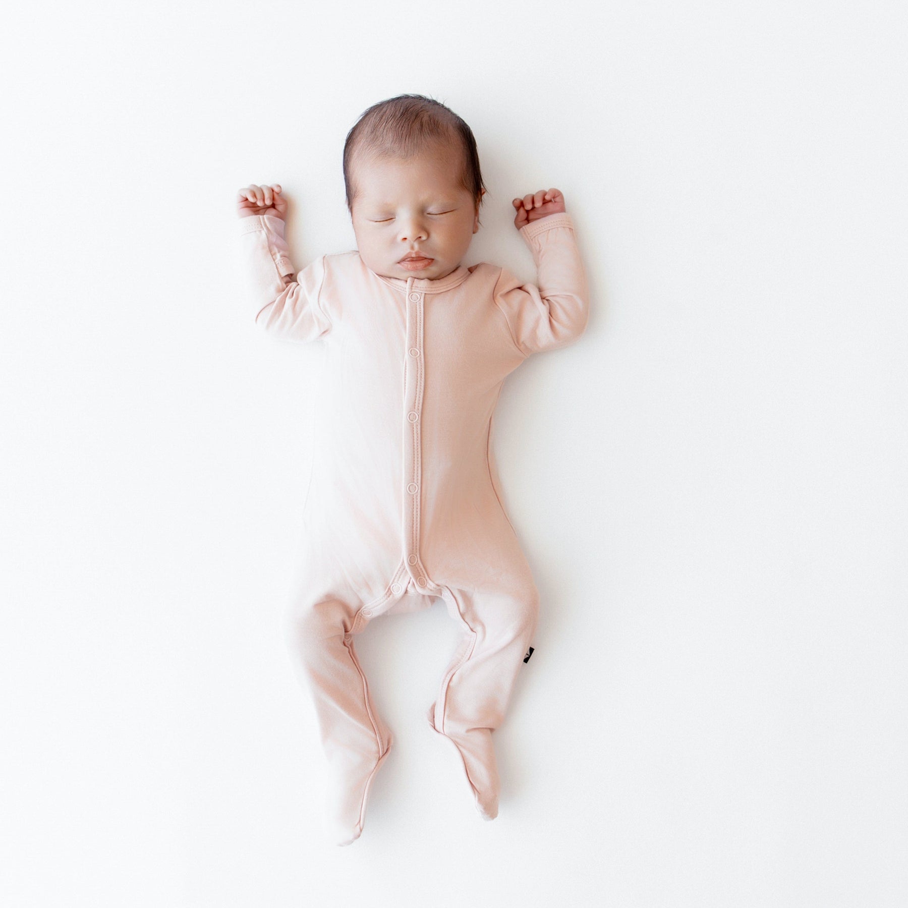 Newborn sleeping on white surface in Kyte Baby Footie in Blush