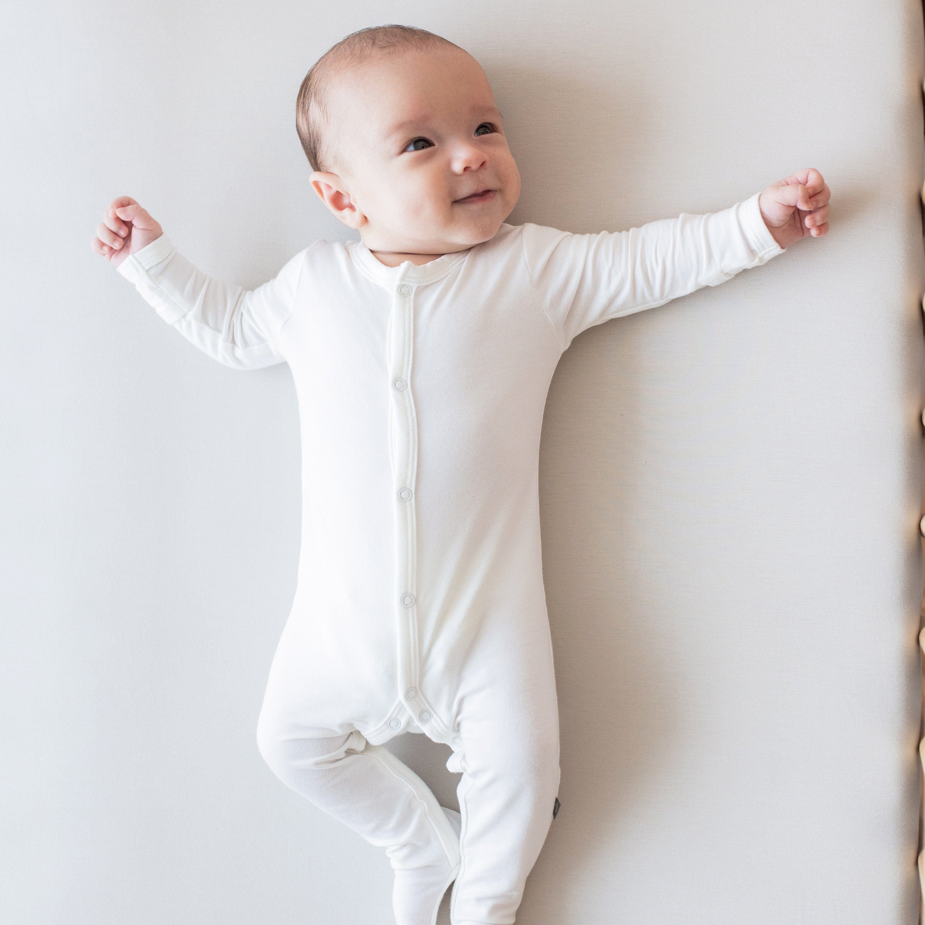 Baby laying on white surface looking towards upper right hand corner, wearing Kyte Baby Snap Footies Footie in Cloud