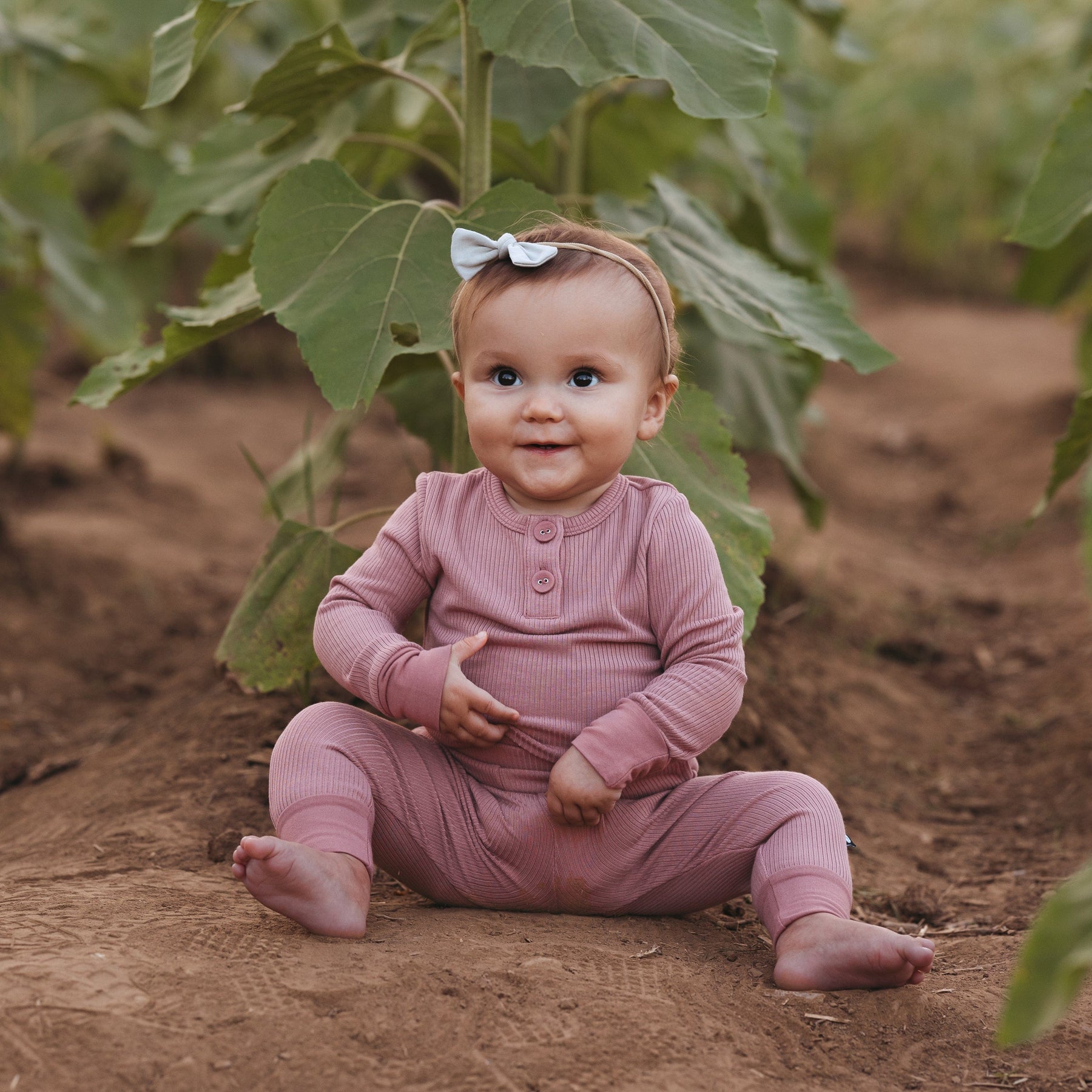 Toddler sitting in front of a large plant on the dirt while wearing a Tiny Nylon Bow in Oat 