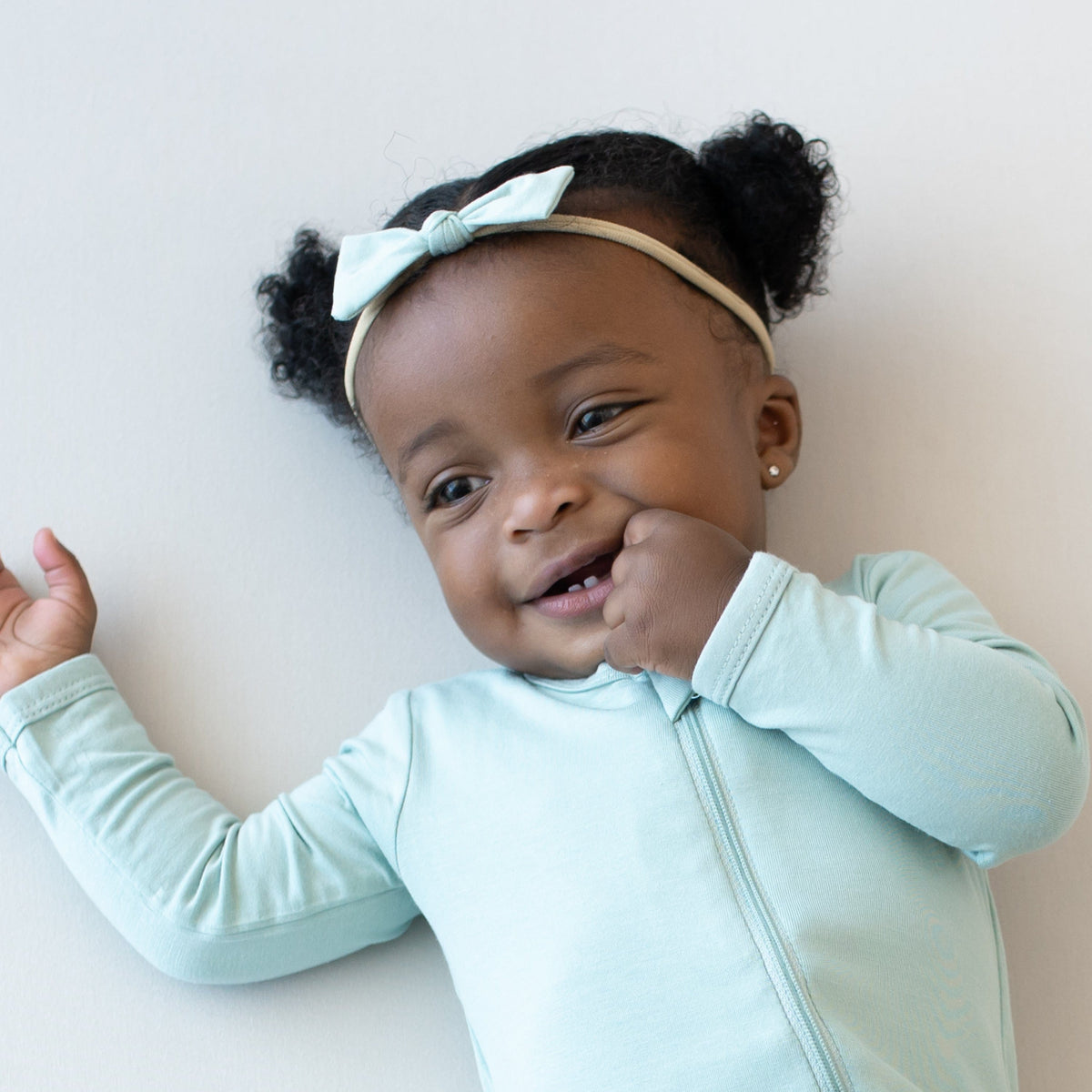 Infant wearing a Kyte Baby Tiny Bow and Zippered Romper in Sage with one hand in mouth