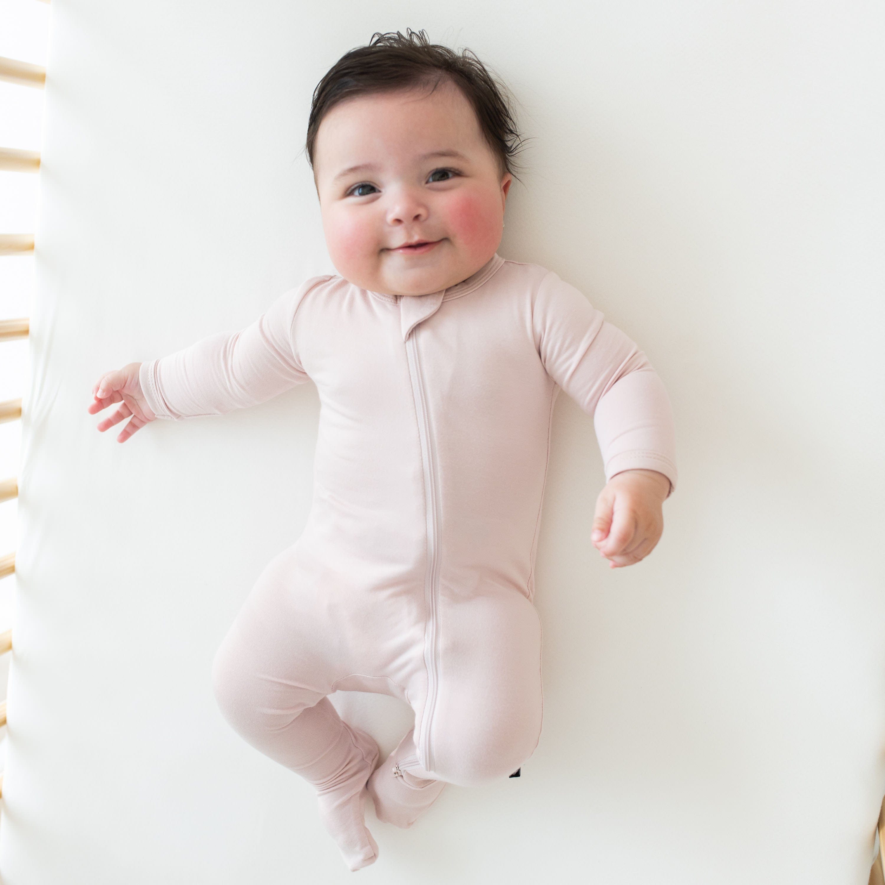Smiling baby laying in a crib while wearing Kyte Baby Zippered Footie in Blush