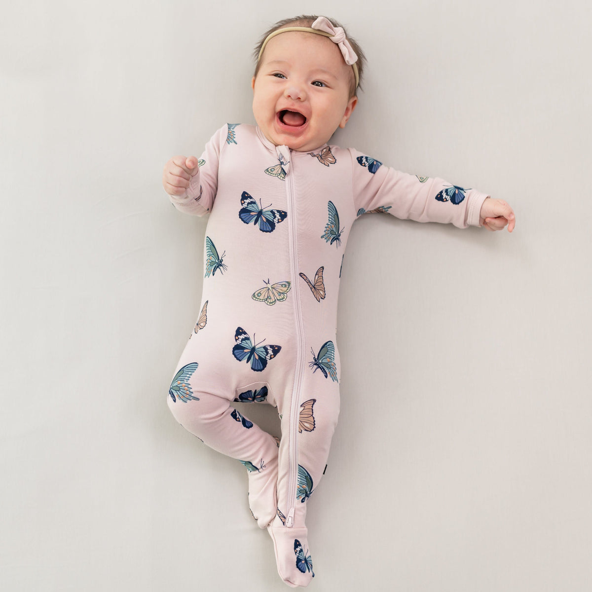 Infant wearing Zippered Footie in Blush Butterfly with pink bow headband