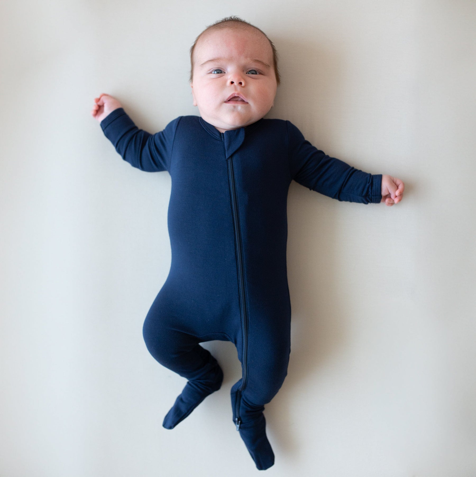 Newborn laying on an off-white surface while wearing a Zippered Footie in Navy