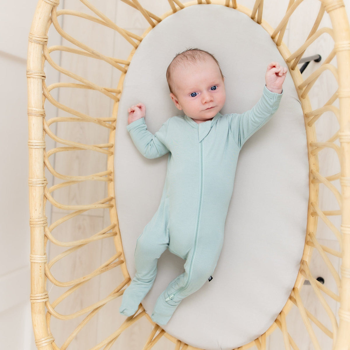 Newborn laying in a rattan bassinet wearing the Zippered Footie in Sage