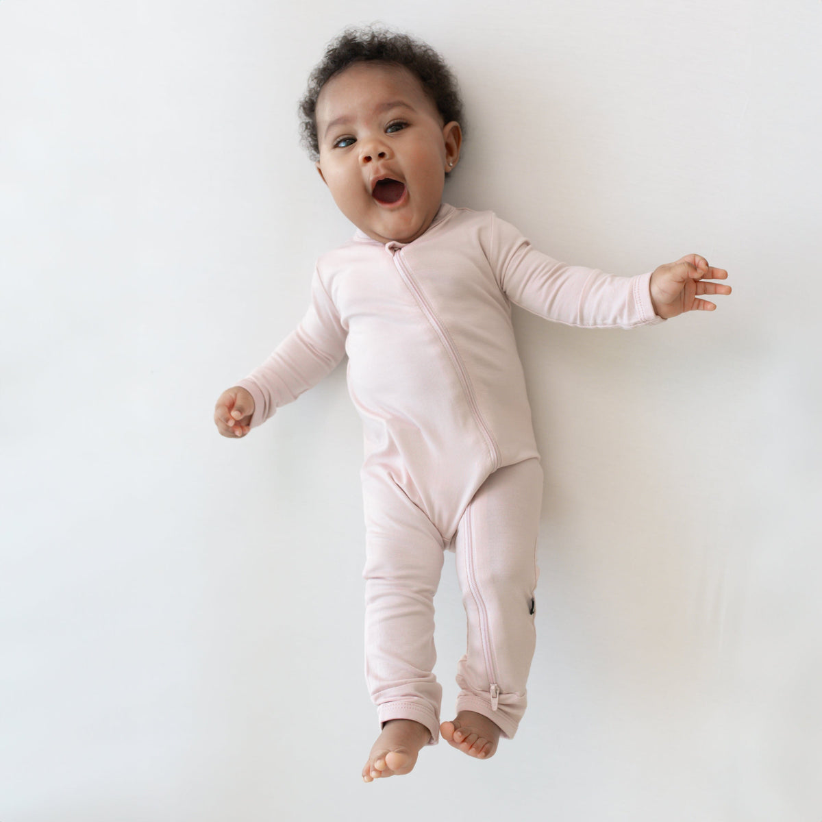 Infant laying on an off-white surface while wearing a Zippered Romper  in Blush