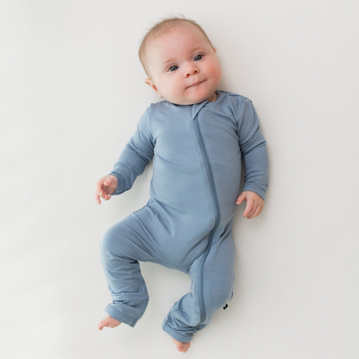 Baby modeling the Kyte Baby  Zippered Romper in Slate while laying on a white surface
