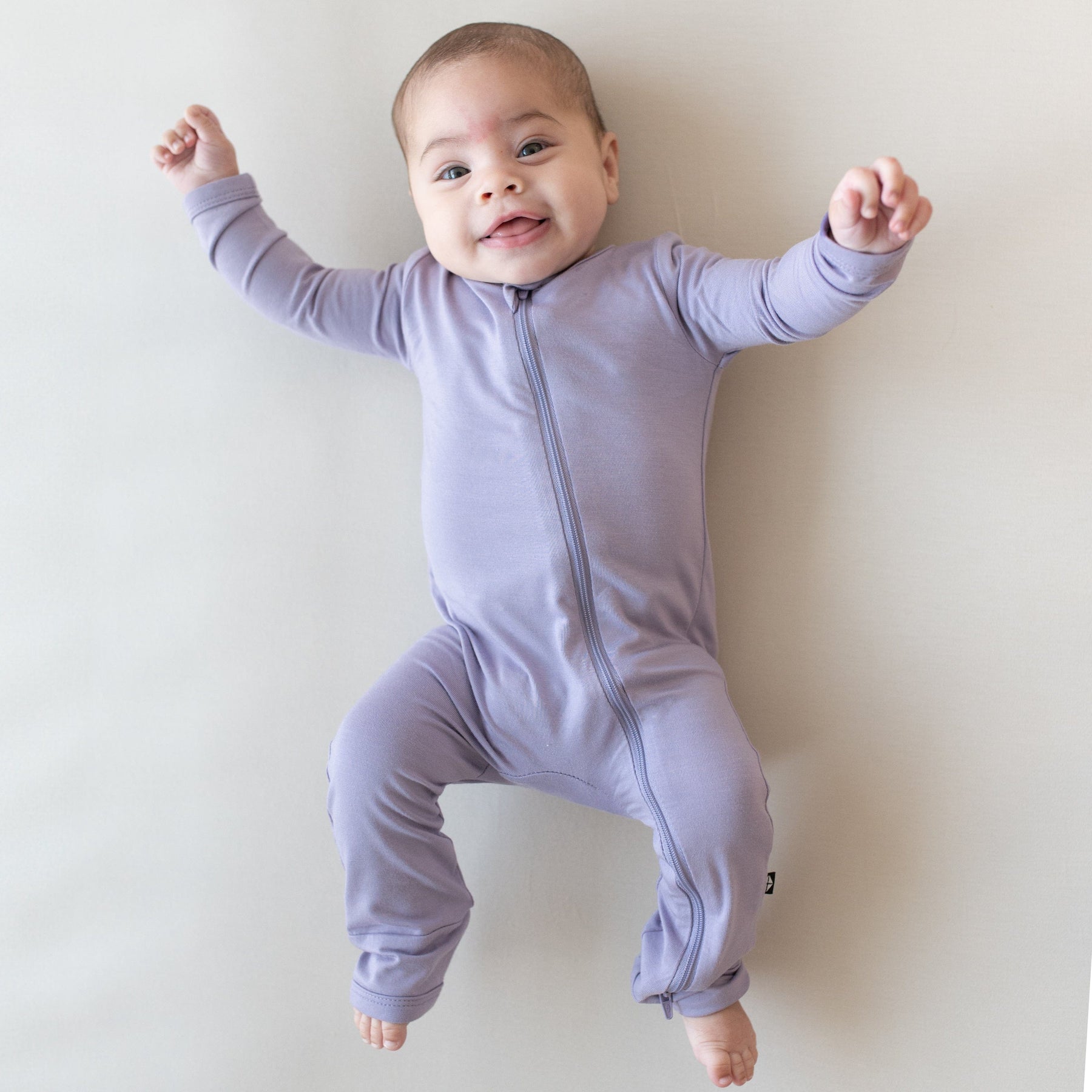 Smiling infant laying on an Oat surface while wearing a Kyte Baby  Zippered Romper in Taro