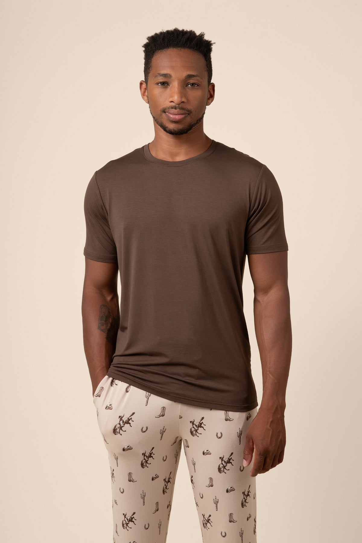 Man wearing a brown t-shirt and western cowboy patterned pants on a beige background