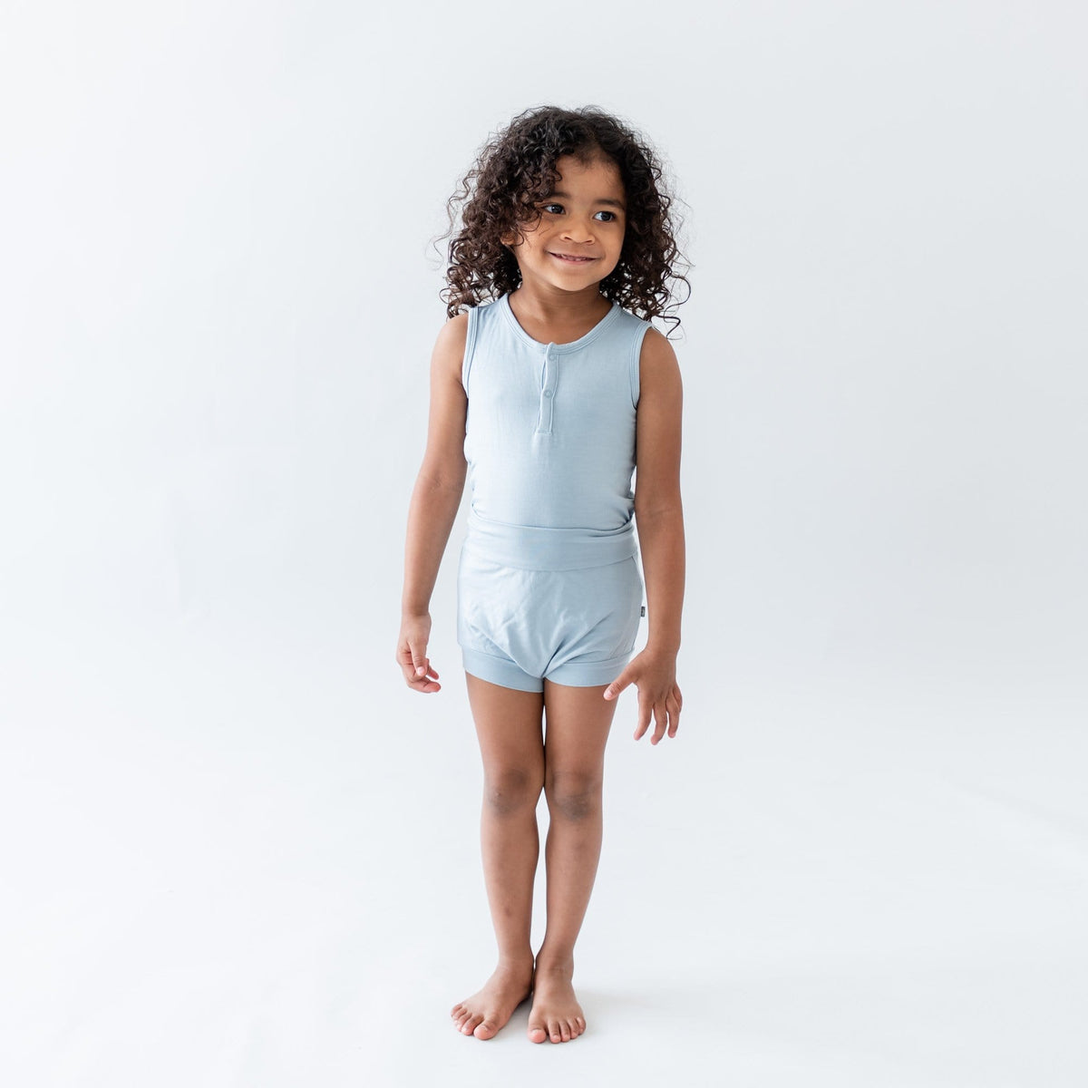 Toddler standing in a white room wearing Kyte Baby Bummies in Fog and matching bodysuit 