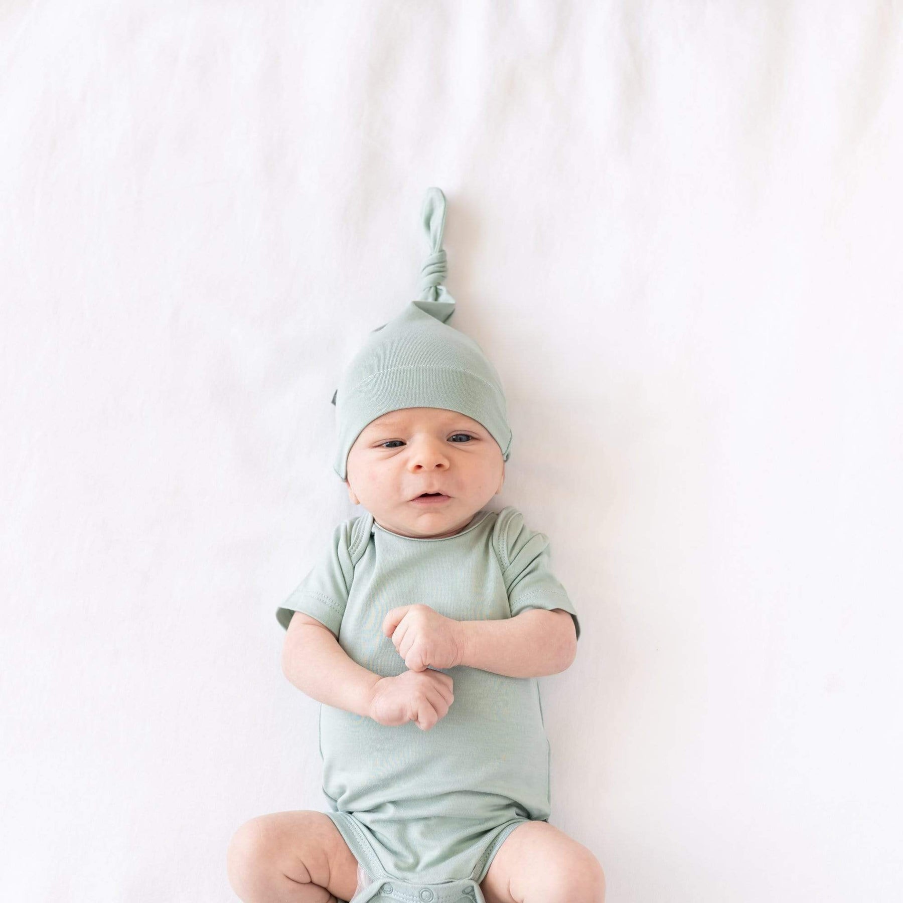 Newborn laying on a crib sheet in Oat while wearing  a Bodysuit and Knotted Cap in Sage 