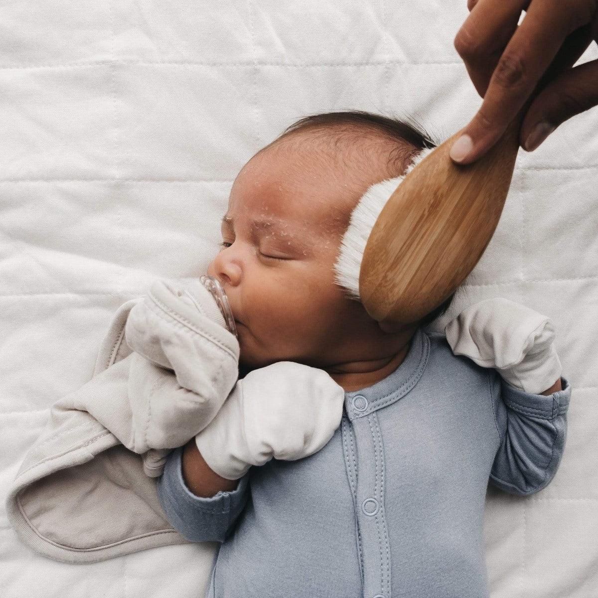Newborn wearing Kyte Baby  Scratch Mittens in Cloud. Baby is sleeping while a soft-bristled brush is used on their head. 