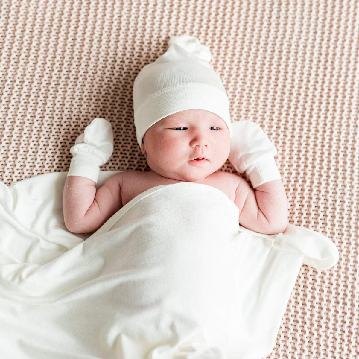 Newborn laying on knitted blanket wearing Kyte Baby Scratch Mittens in Cloud white with matching swaddle and cap