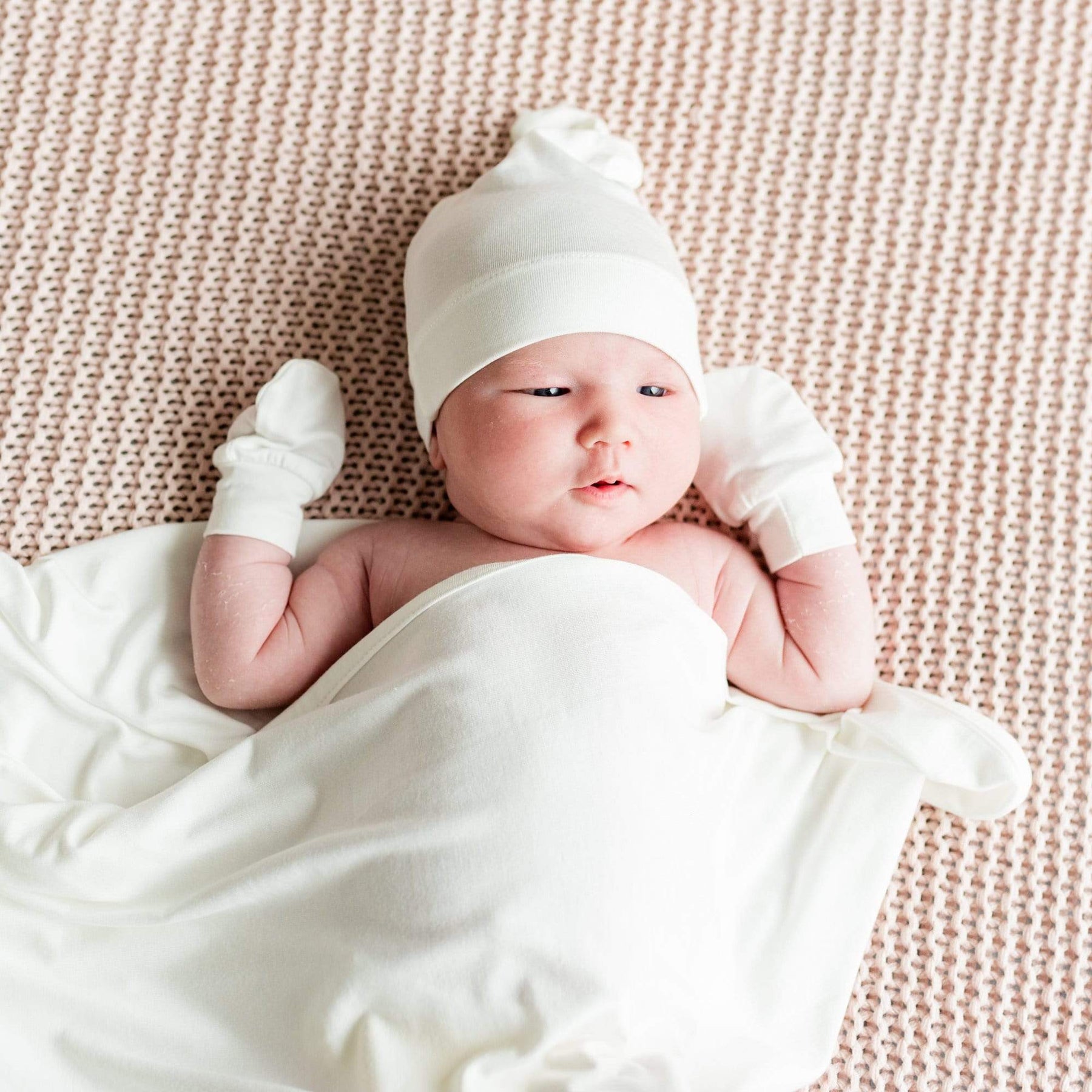 Newborn laying on knitted blanket wearing Kyte Baby Scratch Mittens in Cloud white with matching swaddle and cap