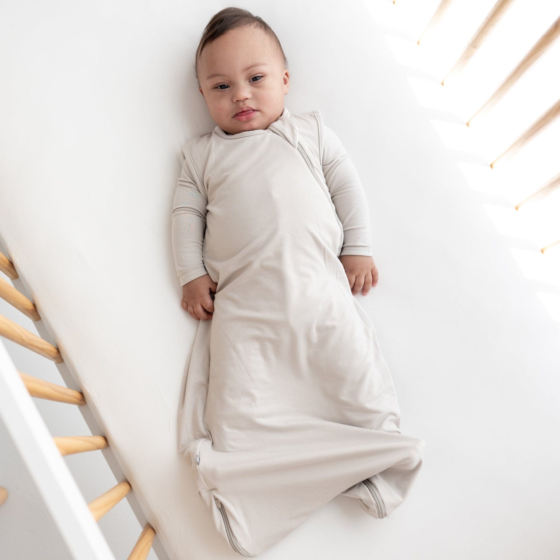 Infant laying in crib while wearing 0.5 Tog Sleep Bag in Oat with a matching long sleeve bodysuit underneath 