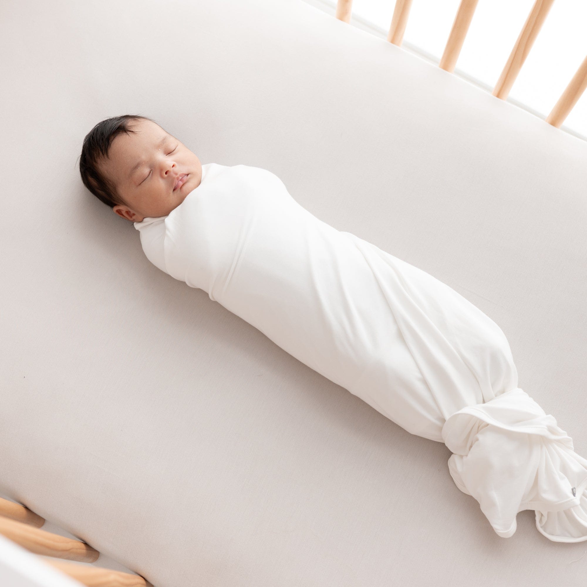 Newborn sleeping in crib while swaddled in Kyte Baby Swaddle Blanket in Cloud