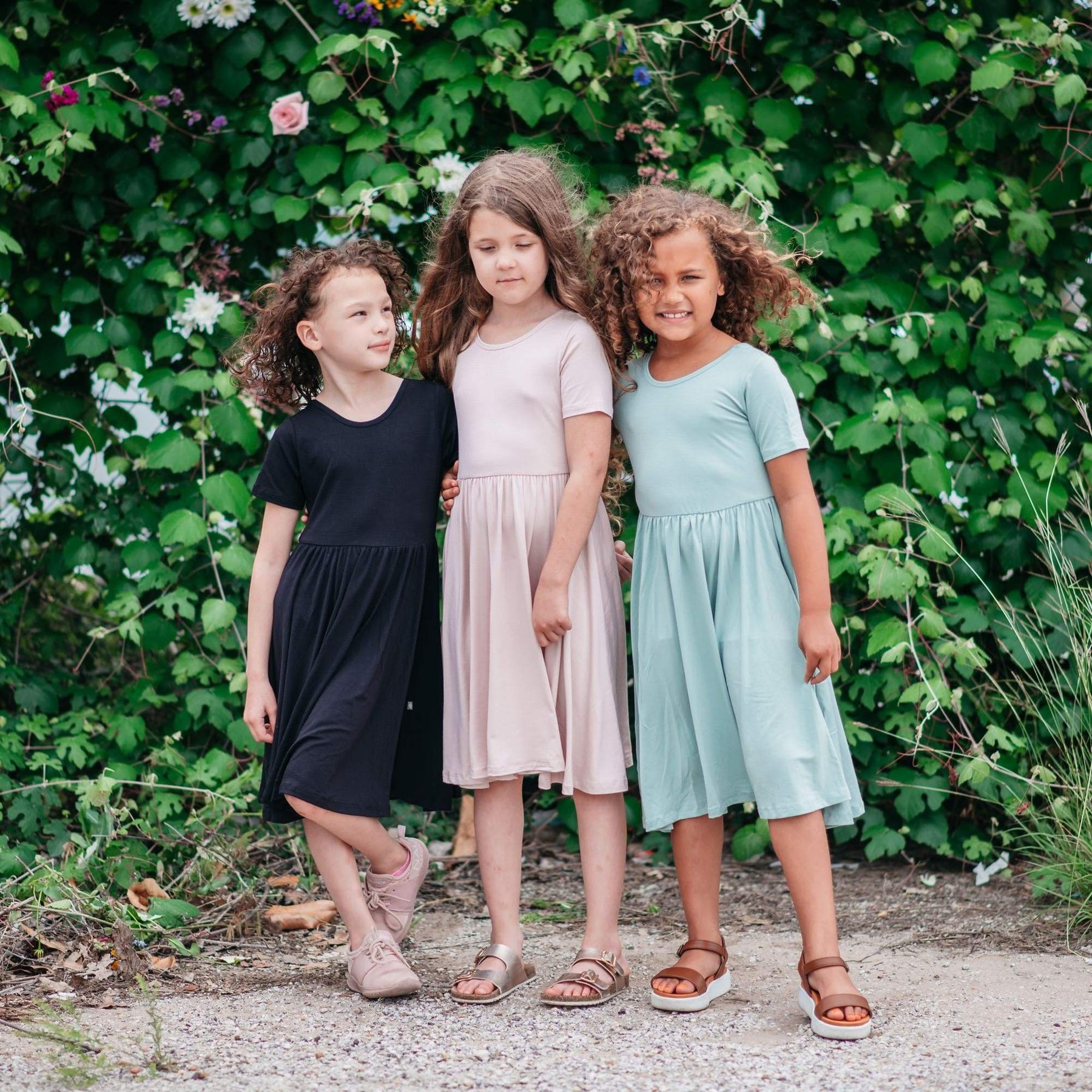 Girls standing next to each other wearing Twirl Dresses in Midnight, Blush and Sage while standing in front of greenery 